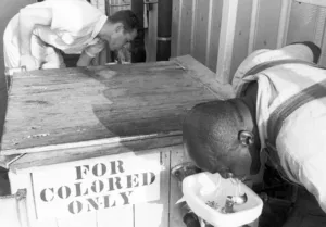 Cena clássica da segregacia racial nos Estados Unidos. Na foto vemos um homem negro bebendo em um bebedouro que deveriam ser usados somente por negros e no outro lado um homem branco bebendo em um bebedouro que só deveria ser utilizado por brancos.